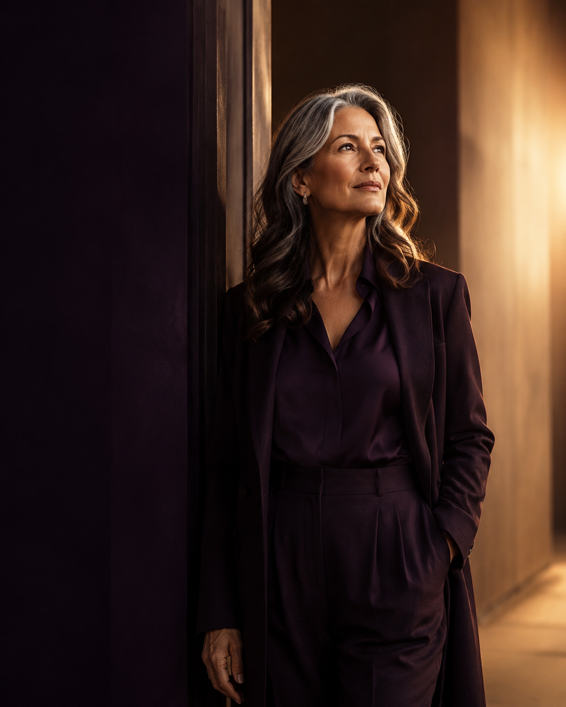 A poised midlife woman standing at a threshold between deep plum shadow and warm golden light